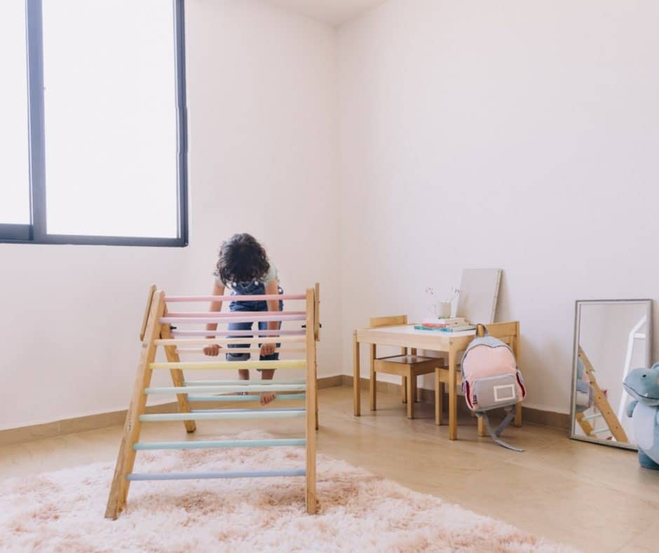 ein heller Raum, links ein Fenster. Hinten in der Ecke ein kleiner Tisch und Stühle. An einem Stuhl hängt ein Rucksack. Auf dem Tisch befindet sich Spielzeug. Im Vordergrund ein heller Teppich auf dem ein PiklerDreieck steht. Auf dem Pikler-Dreieck klettert ein Kind. Spielgruppe Kaltenkirchen, Eltern-Kind-Kurse